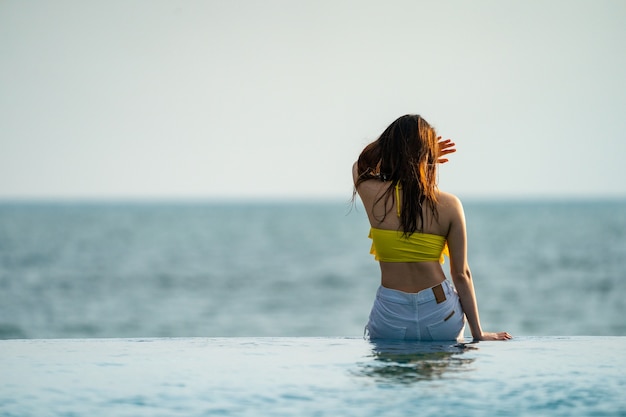 黄色のリラックスと水着のアジアのタイのティーンエイジャーの女性は、タイのリゾートの海のビーチの横にあるインフィニティプールに投稿しています。