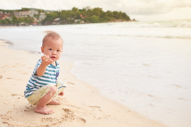 Азиатский малыш ребенок мальчик на пляже с грязными руками, покрытыми мокрым песком, вода на свежем воздухе на летних пляжных каникулах с детьми, сенсорная игра с песком концепции