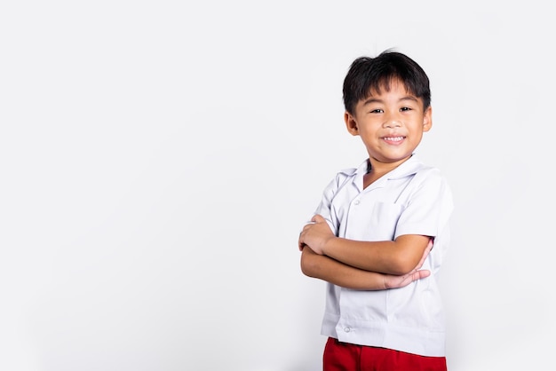 Il sorriso del bambino asiatico felice indossa i pantaloni rossi dell'uniforme tailandese dello studente con le braccia piegate