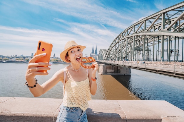 Asian tourist girl taking selfie photo with a pretzel and Cologne cathedral building and bridge over Rhein river on the background Travel and tourism in Germany