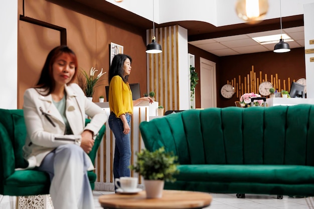 Asian tourist relaxing in hotel lobby. Happy customer sitting in hotel lounge. Female client savoring her coffee inside luxury hotel. Smiling woman enjoying her vacation in elegant hotel.