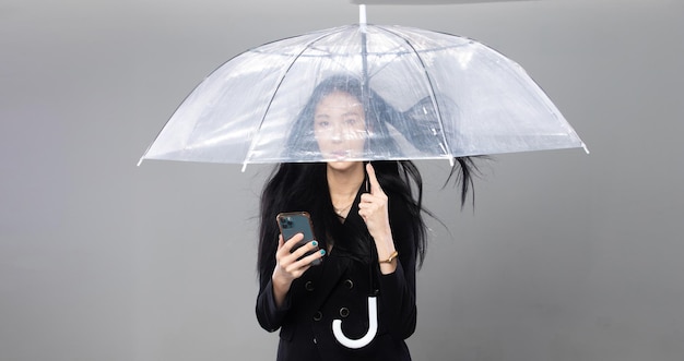 Азиатская трансгендерная женщина с длинными черными прямыми волосами, ветер дует в воздух Женщина держит телефон и зонтик от шторма, чувствуя моду, чувственный сексуальный серый фон, изолированное пространство для копирования