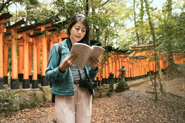 Asian travel woman standing in forest in japanese temple in\
inari shrine in kyoto japan. young girl tourist holding guide book\
looking information carry camera. red gates torii in\
background.