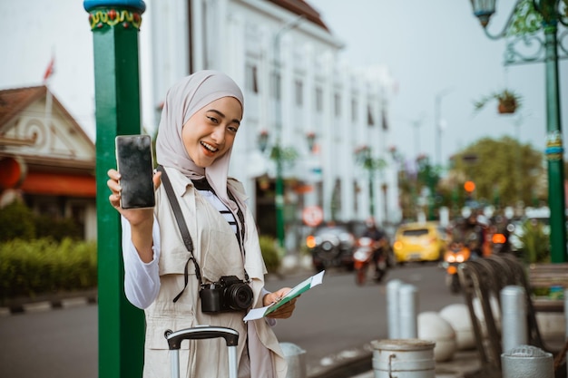 히잡을 쓴 아시아 여행자가 보도에 서서 패스를 가져오는 동안 빈 전화기를 보여줍니다.
