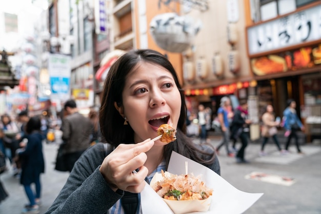 asian visitor eating takoyaki happily. a kind of japanese cuisine. tying oriental food curiously