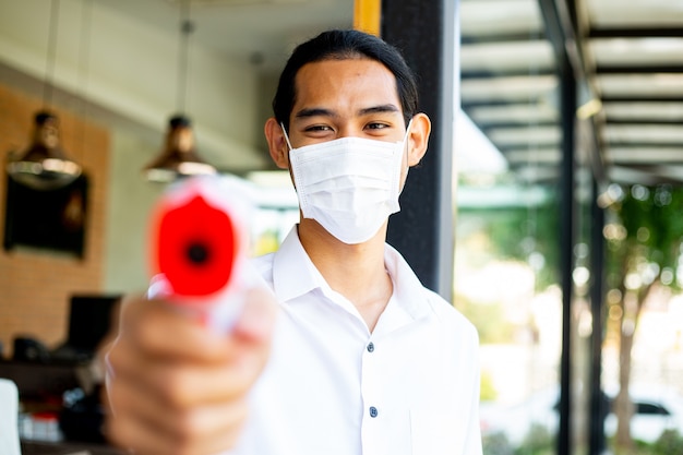 Foto cameriere asiatico che tiene la pistola del termometro per controllare i sintomi della febbre del cliente prima dell'ingresso all'interno del ristorante. prevenire e schermare i malati sani di covid-19.