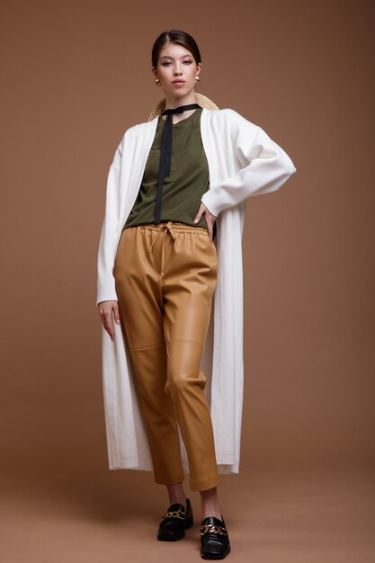 Asian woman in beige leather pants white long cardigan green tshirt belt on brown background