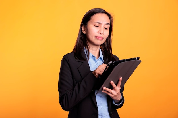 アジア人の女性がタブレット コンピューターで情報を閲覧し、黄色の背景にスタジオで研究プロジェクトに取り組んでいます。クリエイティブな作業にタッチスクリーン付きの電子ガジェットを使用する陽気なモデル。