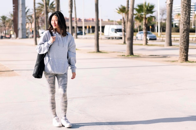 Азиатка в обычной одежде гуляет с ковриком для йоги.