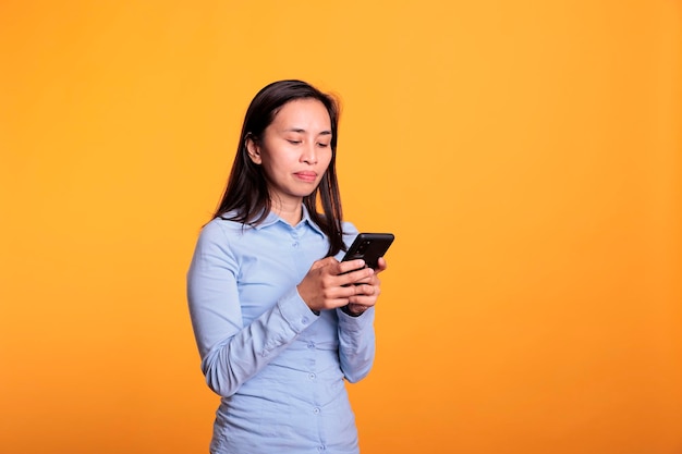 Photo asian woman chatting with remote friend using smartphone, typing message during work break in studio over yellow background. cheerful young adult browsing on social media, searching information