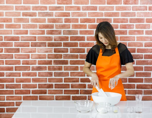 Asian woman chef in orange apron using spatula to stir and mix flour powder in white bowl with water pouring from glass on table near brick wall of home kitchen