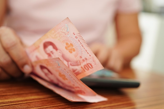 Азиатская женщина подсчитывает деньги в тайских бат и держит в руке бухгалтерский учет инвестиционной экономики