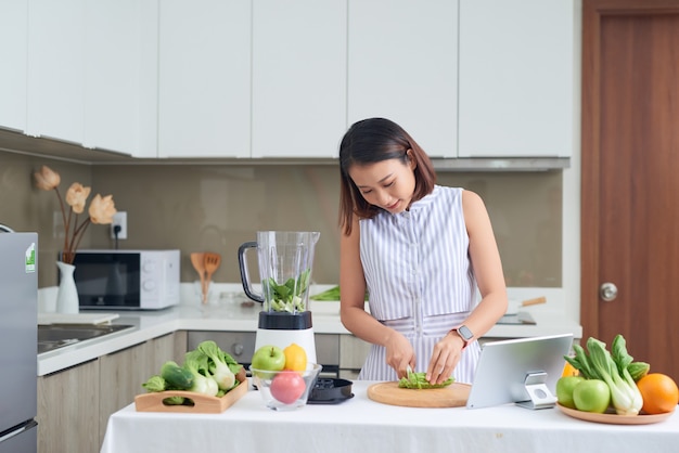 Asian woman cutting vegetables in kitchen with tablet in front of her