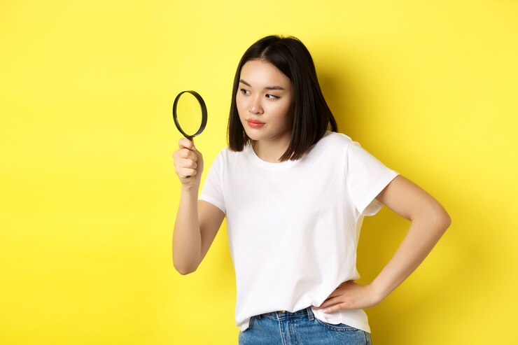 Photo 興味をそそる表情で虫眼鏡を通して見ているアジアの女性探偵は、黄色の背景の上に立って、手がかりを見つけました。