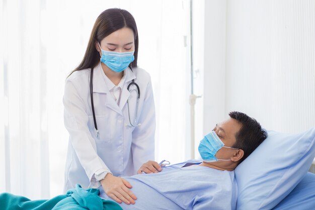 An Asian woman doctor is checking the symptom of a man patient who bed rest in hospital. Both wear a surgical mask to protect Corona virus disease (Covid 19).