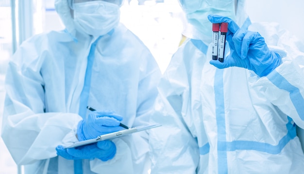 Asian woman doctor in personal protective suit with mask writing on quarantine patient chart, holding test tube with blood sample for screening coronavirus. Coronavirus, covid-19 concept.