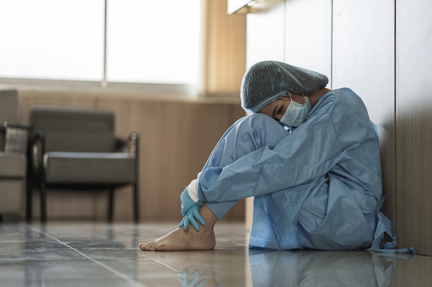 Medico donna asiatica che indossa maschera chirurgica seduta sul pavimento stanco dal lavoro a causa dell'impatto dell'epidemia di pandemia di covid-19, tristezza operatore sanitario donna, concetto medico e sanitario