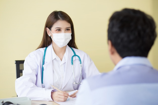 아시아 여성 의사는 의료용 안면 마스크를 착용하고 종이에 남성 환자의 증상을 기록합니다