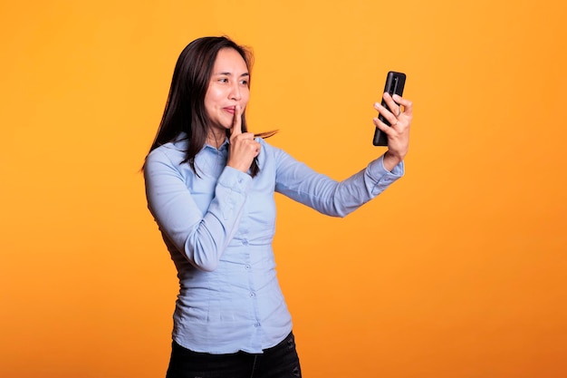 Asian woman doing shh gesture during videocall meeting, having secret discussion with remote friend in studio over yellow background. Cheerful adult enjoying remote conversation during break