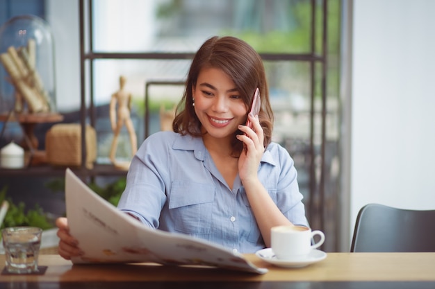 Caffè bevente della donna asiatica nel caffè della caffetteria e per mezzo del telefono cellulare