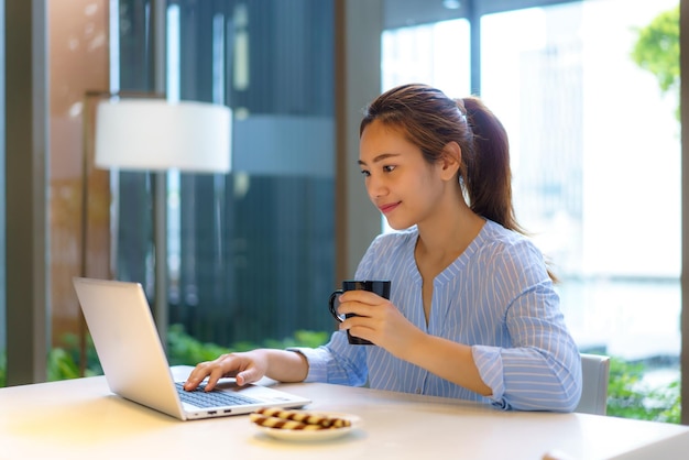 Asian woman drinking coffee in mug and smiling happily while taking a break from work at her computer during work at home in the living room.