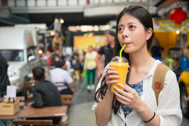 Asian woman drinking the mango juice with a satisfy smile. She is looking at the vendor beside the streets.