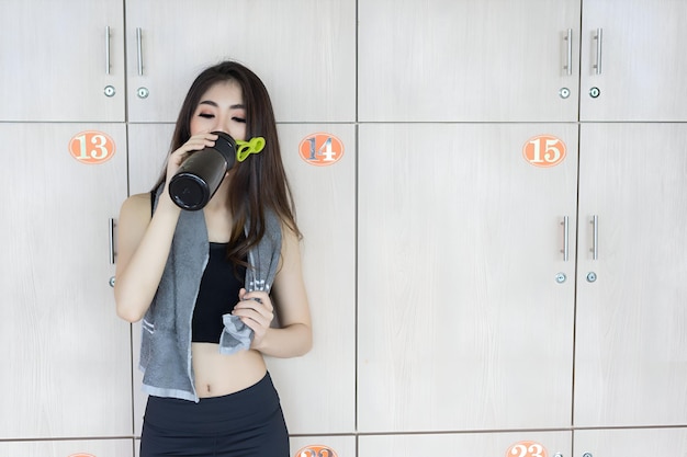 Asian woman drinking protein shake bottle after workout exercise at fitness gym smiling in locker healthy lifestyle bodybuilding