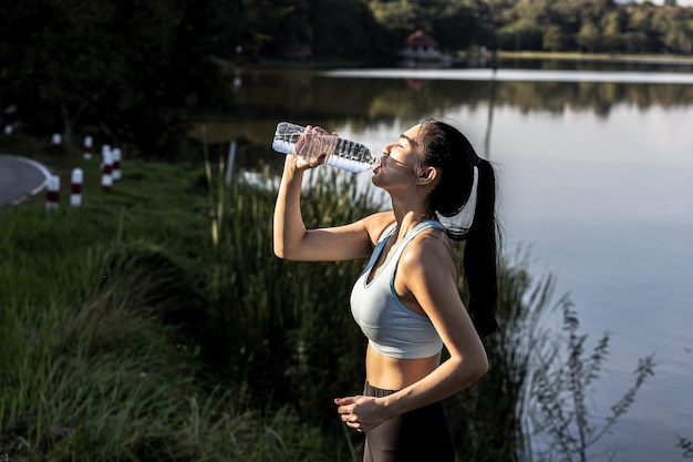 ジョギング後に水を飲むアジア人女性は、朝夕のジョギングに多くの人が訪れる公園でランニングをしており、ランニングが人気のアクティビティです。ジョギングを伴うヘルスケアの概念。