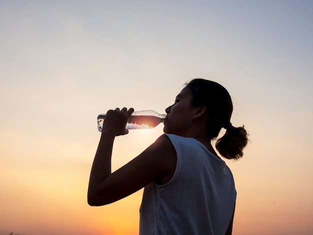 Acqua potabile della donna asiatica dalla bottiglia di plastica dopo l'allenamento che si esercita sull'estate di sera sul bello cielo di tramonto