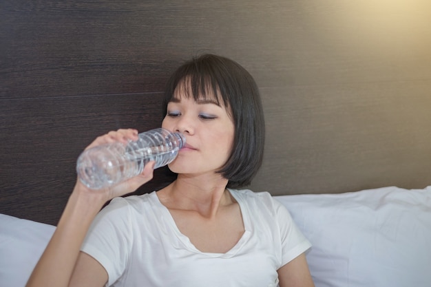 Азиатская женщина пьет воду и сидит в белой кровати дома