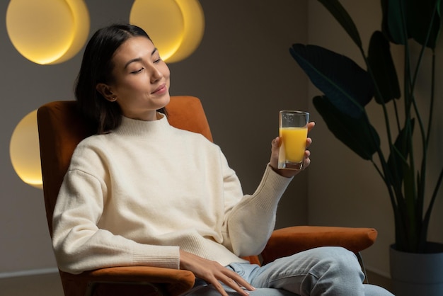 La donna asiatica beve il succo d'arancia da un bicchiere mentre si siede sul divano e si rilassa