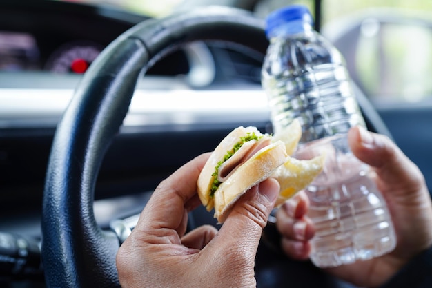 Asian woman driver hold cold water and sandwich bread for eat and drink in car dangerous and risk an accident