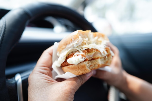 アジアの女性ドライバーは危険な車の中でハンバーガーを持って食べ、事故の危険を冒す