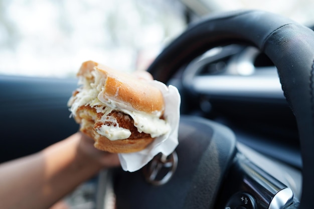 アジア人の女性ドライバーが車の中でハンバーガーを掴んで食べると危険で事故の危険がある