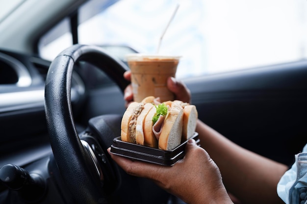 Asian woman driver hold ice coffee cup and sandwich bread for eat and drink in car dangerous and risk an accident