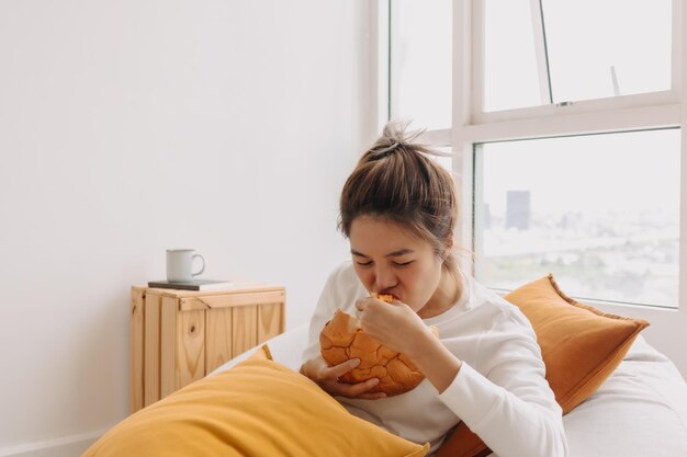 写真 パンとコーヒーの朝食を食べているアジア人女性がリビングで豆袋の上に座っています