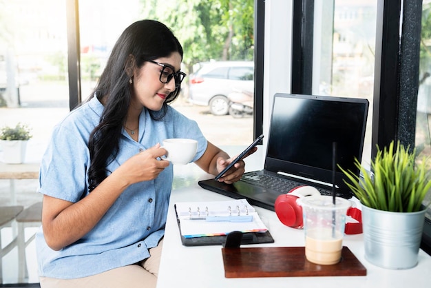 Asian woman in eyeglasses with a laptop and notebook on the table holding cup of coffee while using a mobile phone