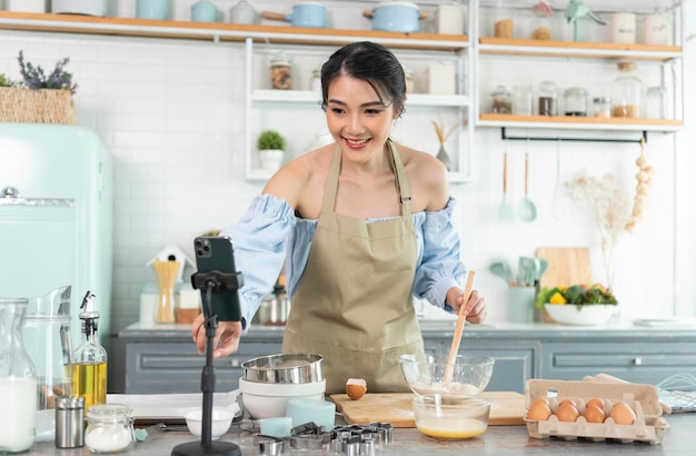 Asian woman food blogger cooking cake in front of smartphone camera while recording vlog video and live streaming at home in kitchen