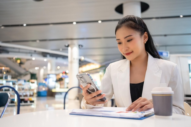 アジアの女性フリーランスカフェで携帯電話で話したりデジタルデバイスやワイヤレステクノロジーでどこからでも働く人々を楽しんでいます若いビジネスウーマンが文書を分析しています