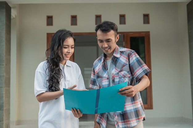 Asian woman hand gesture chatting about home certificate with housing developer while holding house certificate in front of house