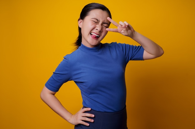 Азиатская женщина счастливая веселая показывая знак мира с двумя пальцами на желтом.