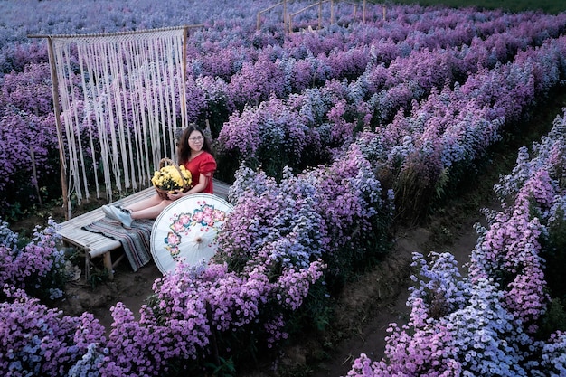 Asian woman happy traveler with red dress hold white umbrella\
enjoying in blooming or purple michaelmas daisy flower field to\
hold a flower basket in the nature garden of in chiang\
mai,thailand
