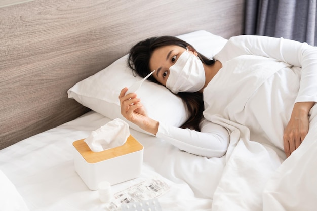 Asian woman has a high fever or infected with COVID19 stay home and self quarantining Ill girl wearing face mask have a headache and cough sleeping on bed Health problems home isolation concept