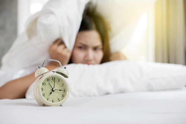 Asian woman hates waking up early in the morning. Sleepy girl looking at alarm clock and trying to h