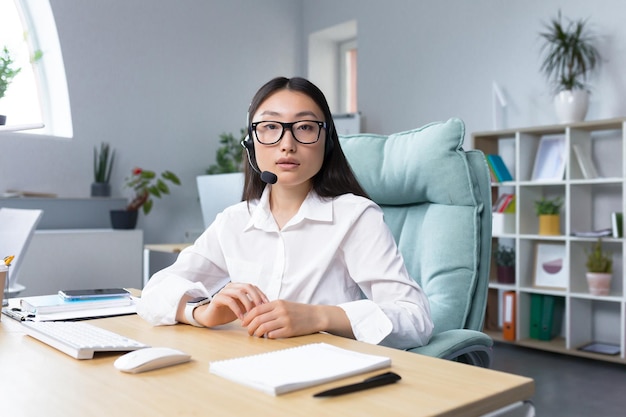 Asian woman in headphones with a microphone sitting in the office at the desk conducting an online