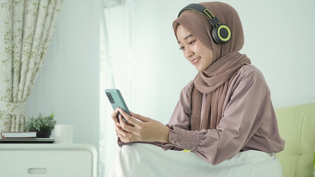 asian woman in hijab is at home listening from her smartphone using headphones