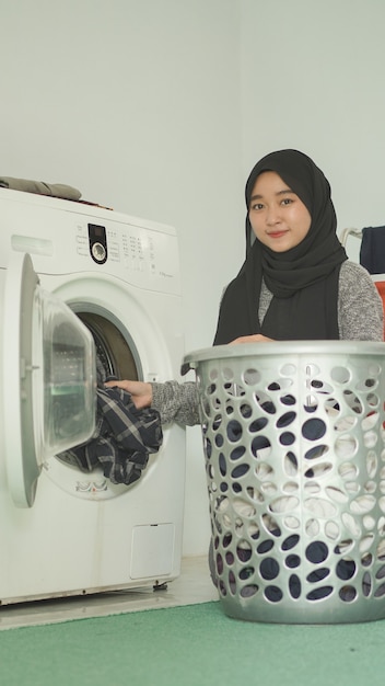 Asian woman in hijab puts dirty clothes in washing machine at home