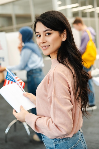 Asian woman holding American flag and ballot paper looking at camera standing at polling station