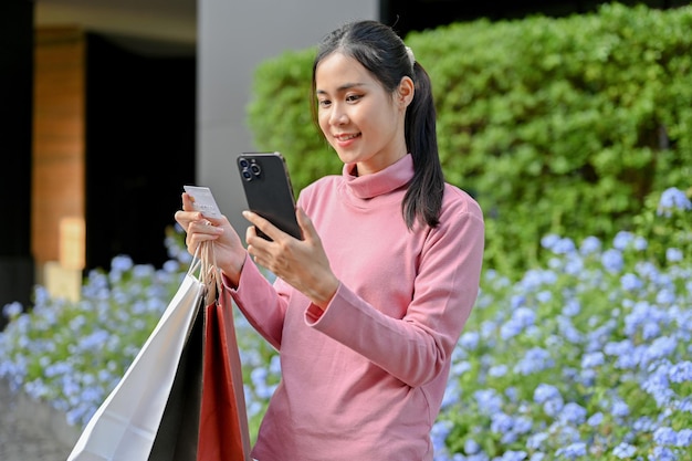 Asian woman holding credit card using smartphone carrying shopping bags