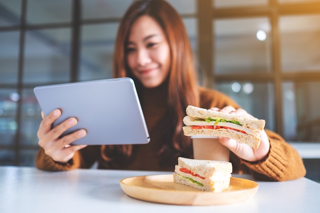 An asian woman holding and eating whole wheat sandwich while using tablet pc